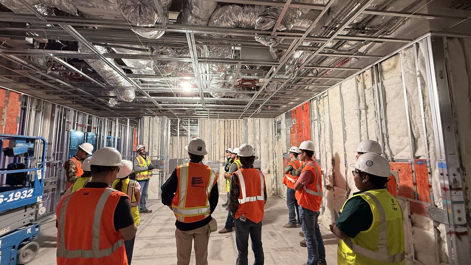 Students listening to a site professional inside a building with exposed metal framing, insulation, and ceiling ducts