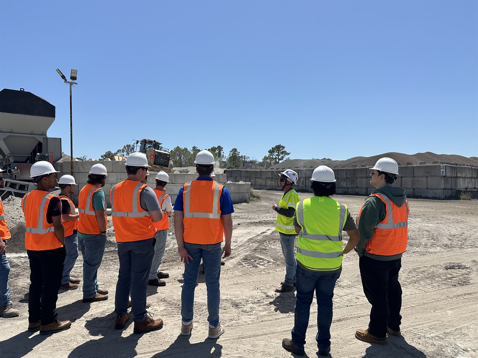 Students listening to a construction professional beside aggregate stockpiles, concrete barriers, and construction equipment