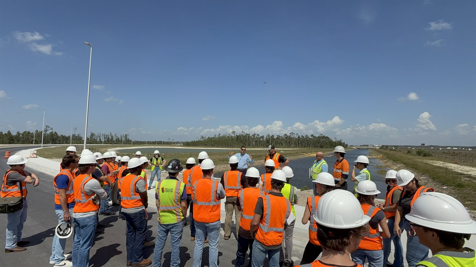 Students in hard hats and safety vests listening to construction professionals beside a roadway and drainage area