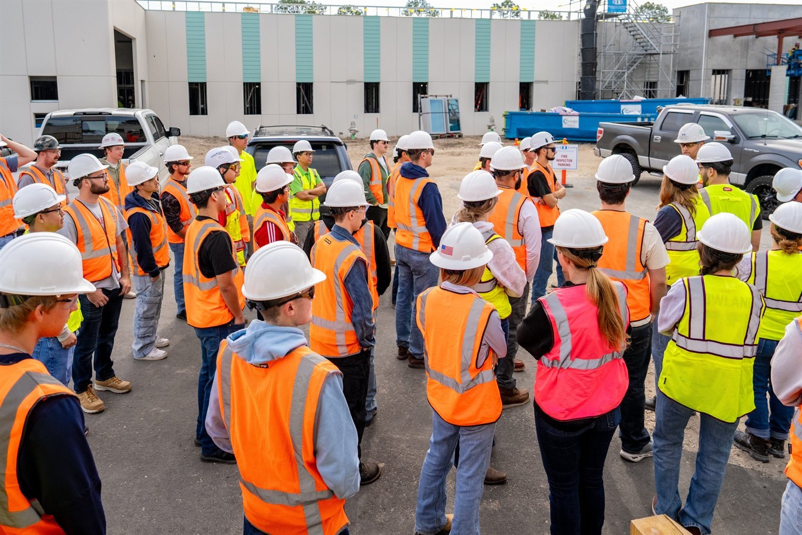 Students in hard hats and safety vests listening to a construction professional beside a building under construction