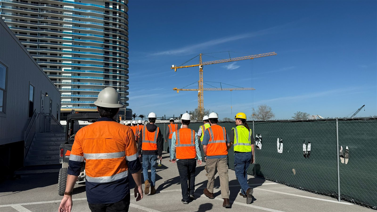 Students walking through a construction site beside a high-rise building and tower cranes
